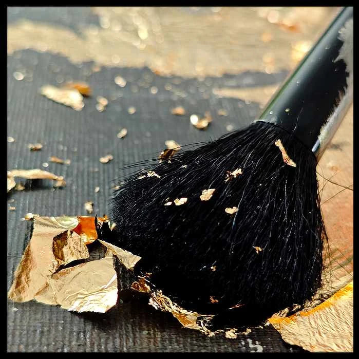 Gold brushstrokes dance on smooth canvas, revealing textured strokes of a black paintbrush. Luxurious abstract art with sophisticated elegance.